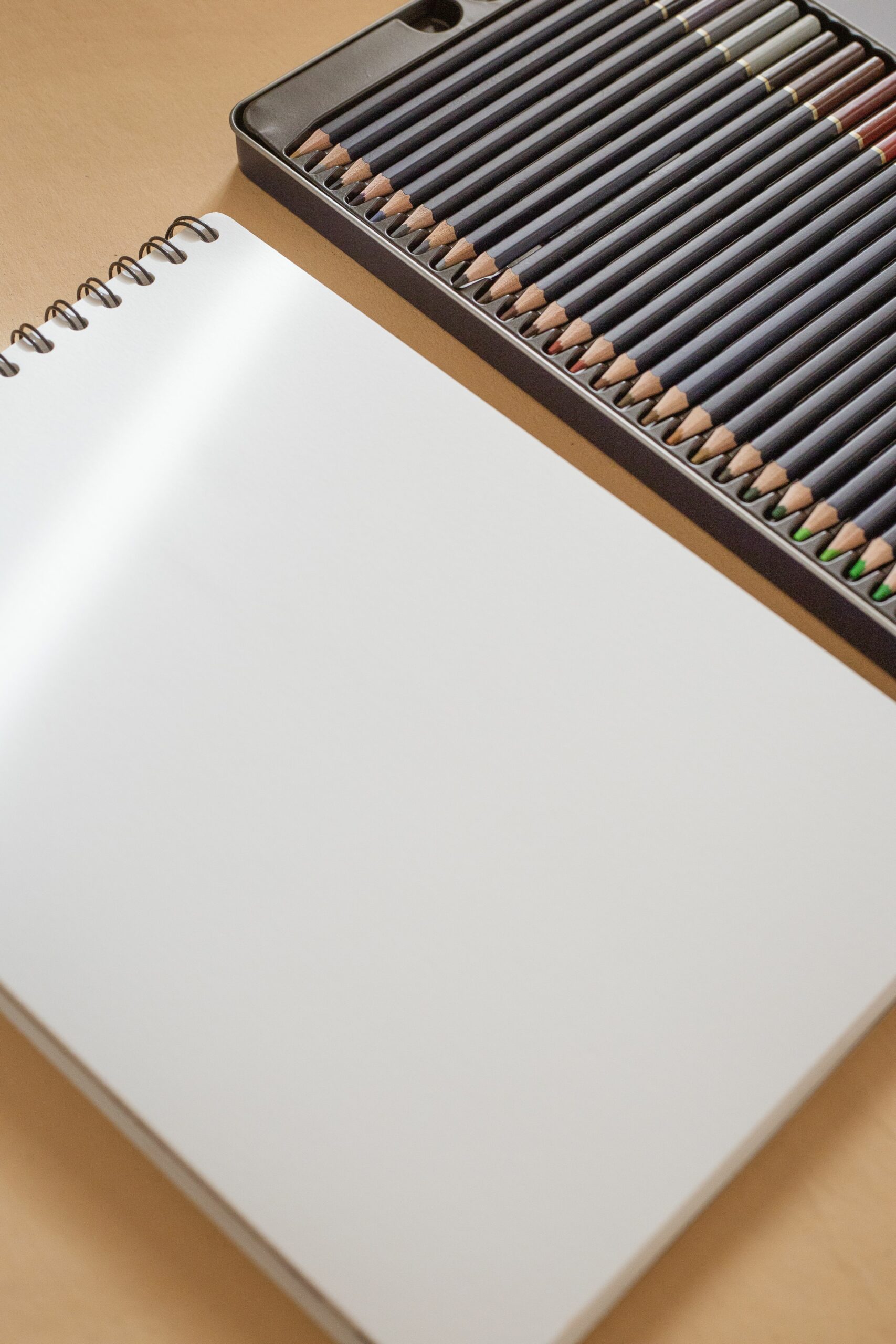 Colorful pencils arranged beside a blank spiral-bound sketchbook on a wooden surface.