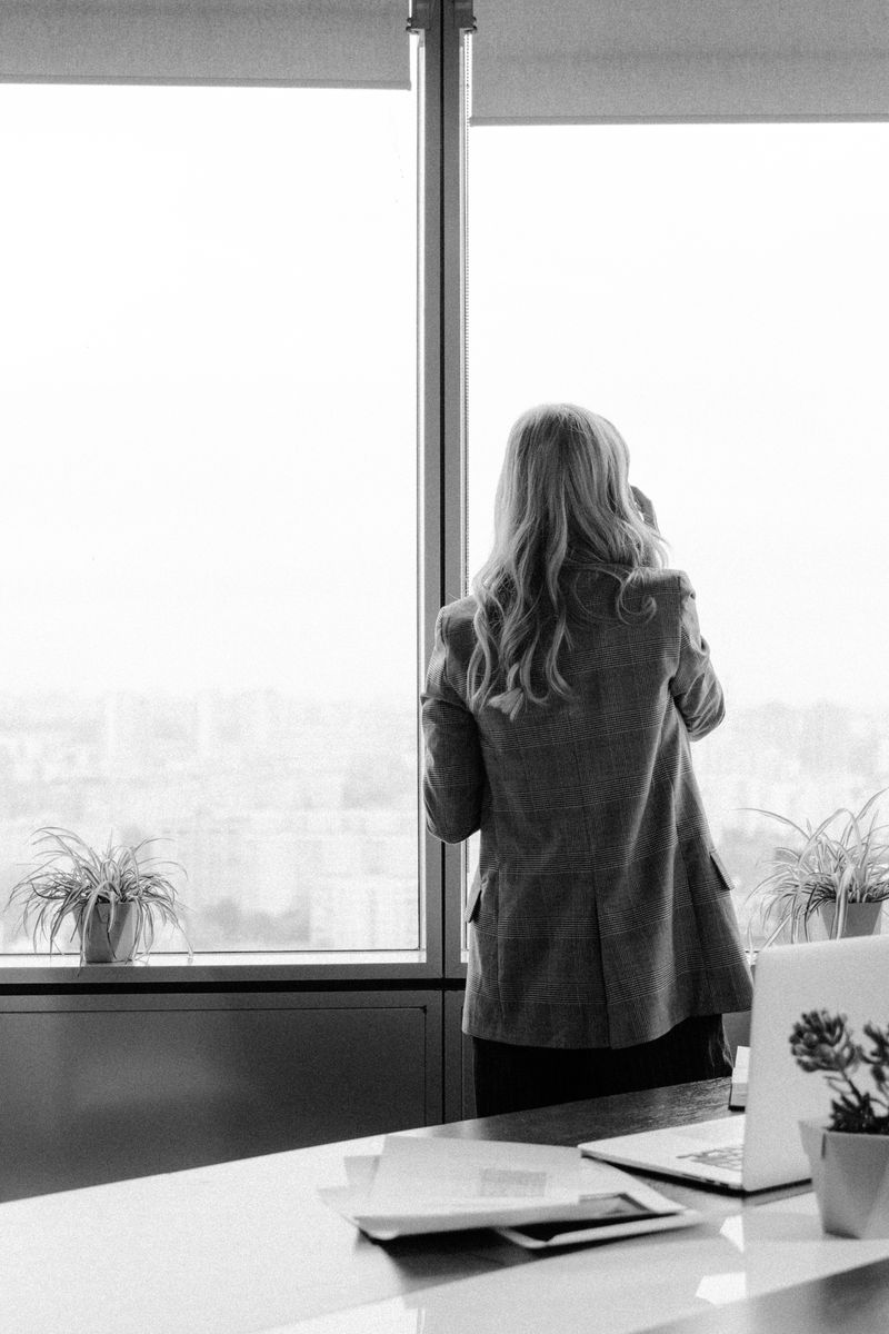 A female executive gazes out the window in a modern office setting, in grayscale photography.